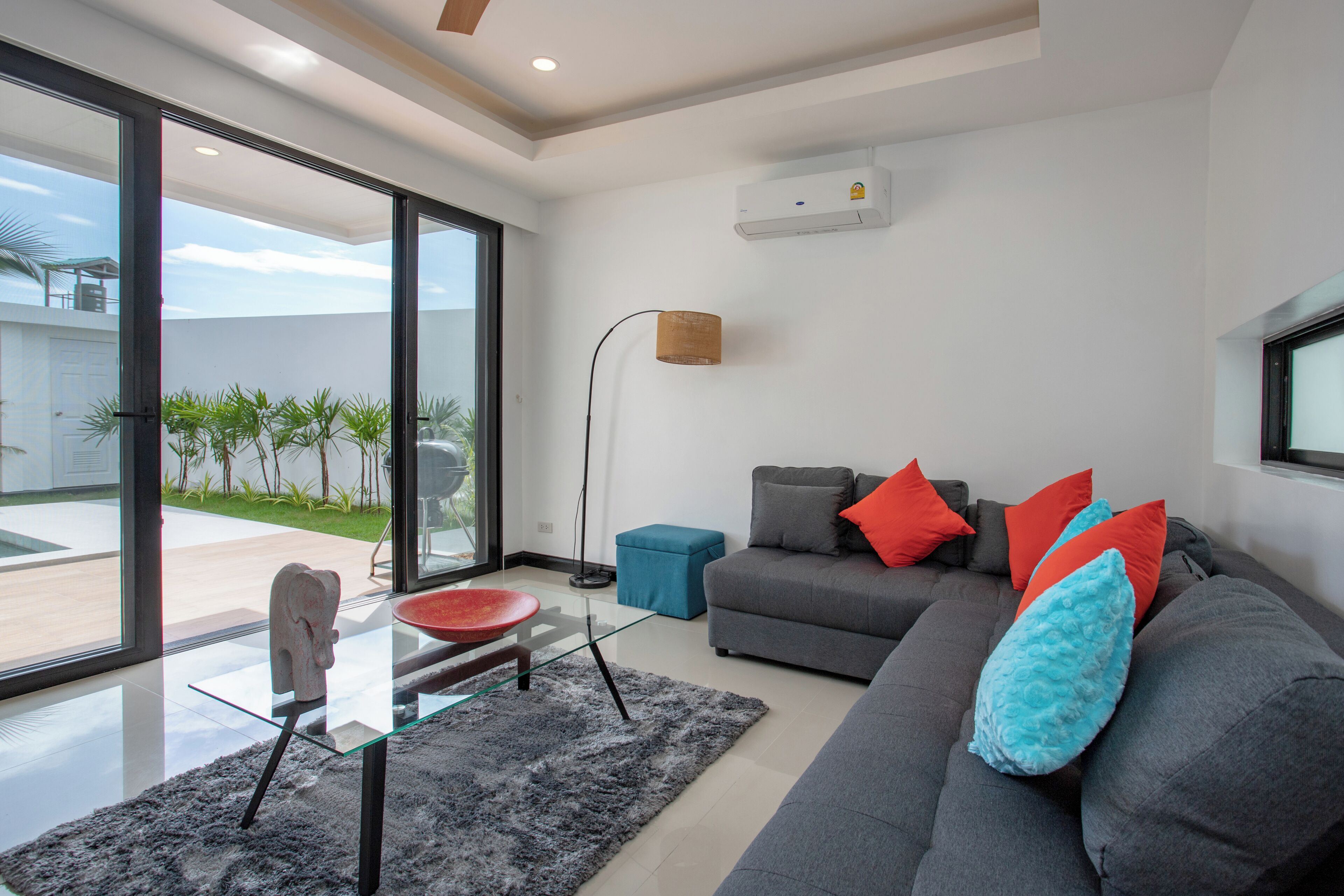 A modern living room with a grey sofa, red and blue cushions, a glass coffee table, and a floor lamp.