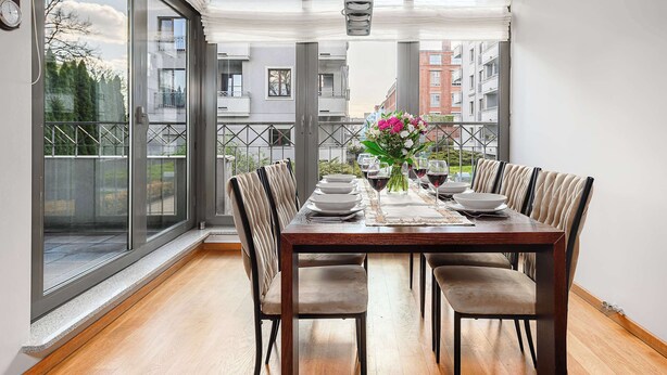 A dining area with a wooden table set for four, large windows, and a view of buildings. - Lumina stylish apartment with parking (Lódz)