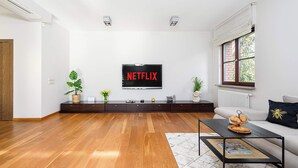 A modern living room with a wooden floor, a sofa, a coffee table, and a TV mounted on the wall displaying 'NETFLIX'. - Lumina stylish apartment with parking (Lódz)
