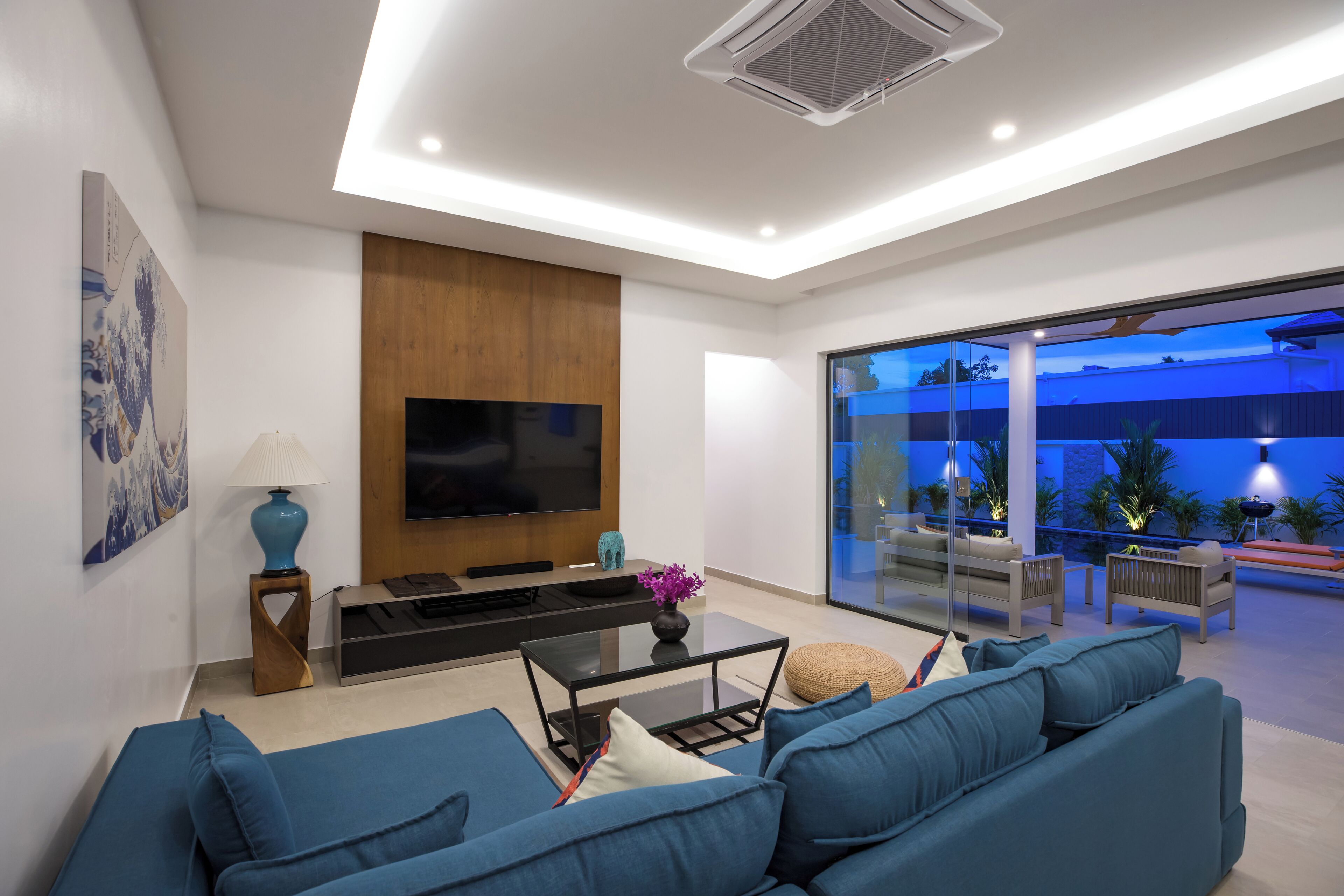 A modern living room with a large flat-screen TV, blue sofas, a coffee table, and a glass door leading to an outdoor patio area.