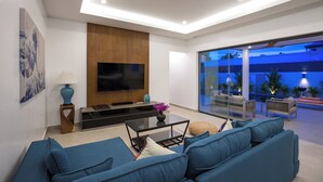 A modern living room with a large flat-screen TV, blue sofas, a coffee table, and a glass door leading to an outdoor patio area. - Luxurious 3 bedrooms pool villa at rawai (Tambon Rawai)
