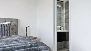 Une chambre à coucher avec une tête de lit capitonnée, une couverture grise moelleuse, une table de chevet et une salle de bain moderne dotée d’une douche et d’un lavabo en marbre.