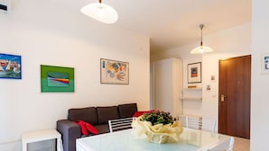A modern living room with a glass-top dining table, white chairs, a brown sofa, and a small white side table. - Nice apartment for 5 guests with WIFI, A/C, TV, terrace and pets allowed (San Bartolomeo Al Mare)