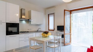 A modern kitchen with white cabinets, a gas stove, and a built-in oven. There is a dining table with chairs and a vase of flowers. A television is mounted on the wall, and a window with curtains is visible. - Nice apartment for 5 guests with WIFI, A/C, TV, terrace and pets allowed (San Bartolomeo Al Mare)