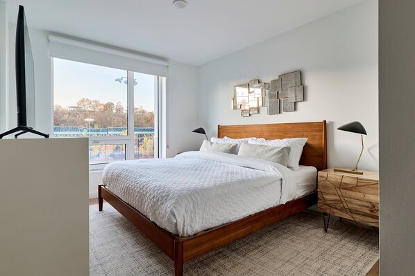A bedroom with a large window, a wooden bed, a nightstand, and wall art.