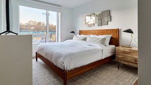 A bedroom with a large window, a wooden bed, a nightstand, and wall art. - Modern 2br-newly Built-near NYC (Hoboken)