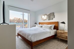 A bedroom with a large window, a wooden bed, a nightstand, and wall art. - Modern 2br-newly Built-near NYC (Hoboken)