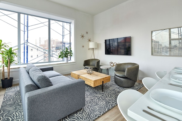 Un salon moderne avec un canapé gris, une table basse en bois et un tapis à motifs zébrés. On y trouve deux fauteuils, une lampe et une table à manger avec des assiettes blanches.