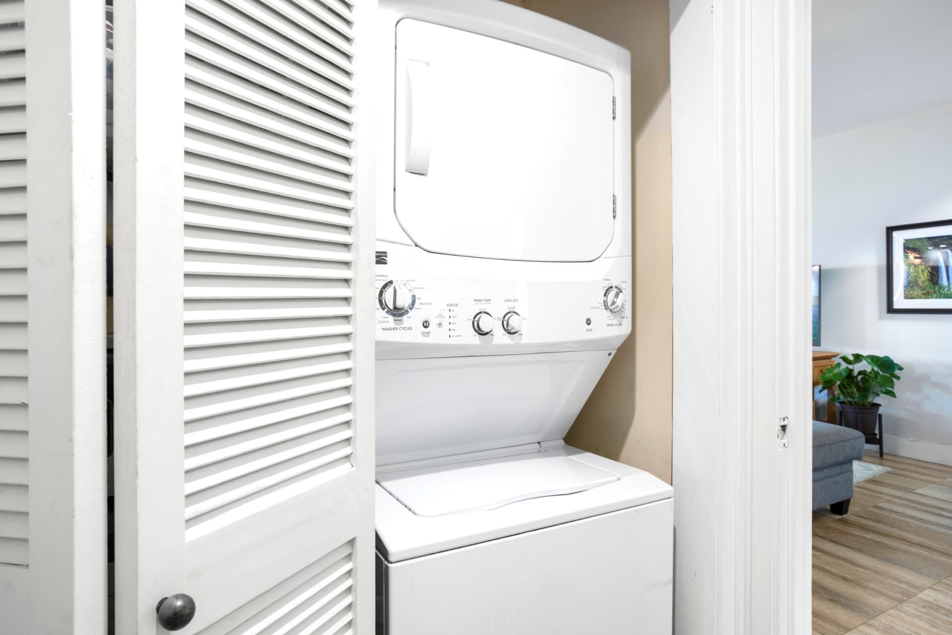 A compact laundry room with a front-loading washer and dryer, a control panel with various settings, and a door leading to a living area with a couch and a plant.