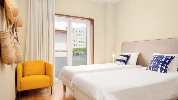Habitación de hotel con dos camas, un sillón amarillo y vista a un edificio a través de la ventana.