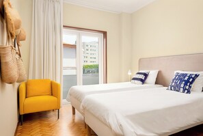 Ett hotellrum med två sängar, en gul fåtölj och utsikt över en byggnad genom fönstret.