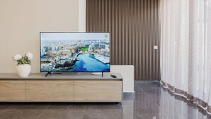 Ein modernes Wohnzimmer mit einem Flachbildfernseher auf einer Holzkonsole, einer Topfpflanze und Blick auf eine Stadtansicht durch das Fenster.