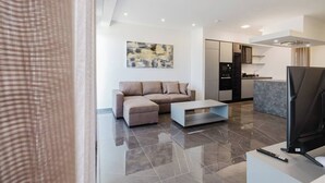 A modern living room with a sofa, a coffee table, a TV, and a kitchen area in the background. - Enjoy a stylish experience at this centrally-located place. Enjoy easy access to Qawra promenade only seconds away from parks and the aquarium.  Modern and stylish, this apartment in Qawra is the ideal place to spend a holiday. (San Pawl il-Baħar)