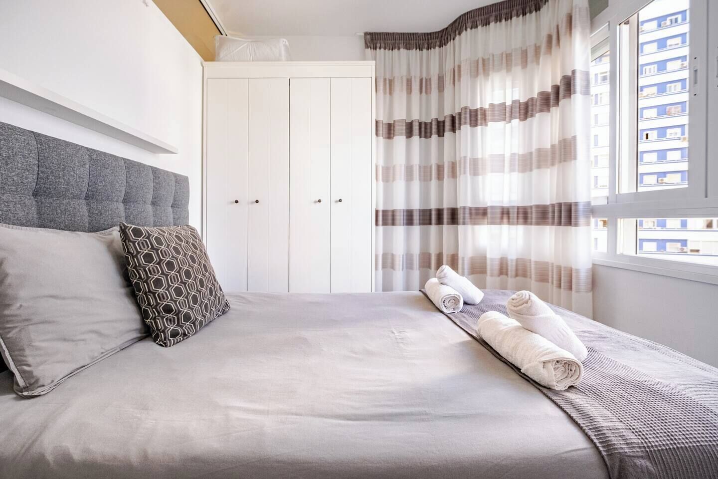 Un lit bien fait, avec une tête de lit grise, un coussin à motifs et des serviettes enroulées posées sur un couvre-lit blanc.