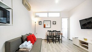 En moderne stue med en grå sofa, røde puder, et spisebord med stole, et fladskærms-tv og et lille tv på et skab.