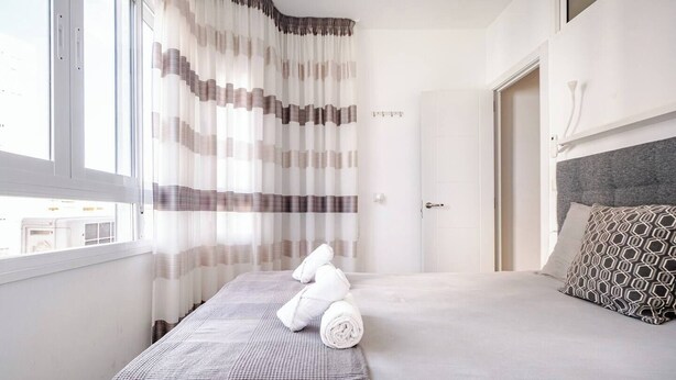 Et hotelværelse med seng, gardiner foran et vindue, en dør og et sengebord med pude.