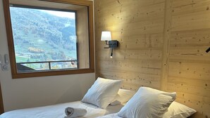 A bedroom with two beds, a wooden wall, a window with a view, and a bedside lamp.