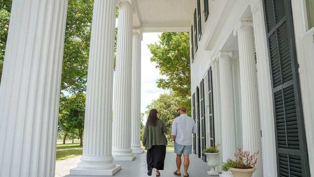 A covered walkway with white columns and shutters, leading to a building.
