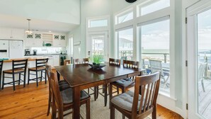 A spacious dining area with a wooden table and chairs, a clock on the wall, and large windows offering a view of the outdoors. - Ocean Breeze - 2nd Row, Best Sunrises, Surf Sounds (Freeport)