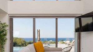 A modern living room with a large window, a sofa, a glass coffee table, and a television. - Kassiani Villa | Theo Luxury Villas With Sea View (Agia Marina)