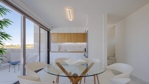 Un espace repas moderne avec une table en verre et des chaises blanches, un meuble en bois au-dessus, et une vue sur l’extérieur.