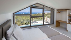 Une chambre dotée d’une grande lucarne, d’un lit, d’une armoire en bois et offrant une vue sur la mer et la verdure.