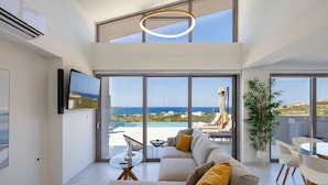 Un salon moderne doté d’un grand canapé, d’une table à manger et offrant une vue sur la mer grâce à de grandes fenêtres.