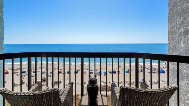A balcony with wicker chairs overlooking a beach with umbrellas and people.