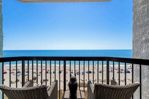 Un balcon avec des fauteuils en osier donnant sur une plage parsemée de parasols et où se trouvent des gens.