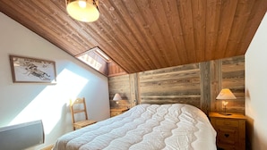 Un dormitorio con techo de madera, una cama con colcha, dos mesitas de noche con lámparas, una silla y una ventana con luz natural.