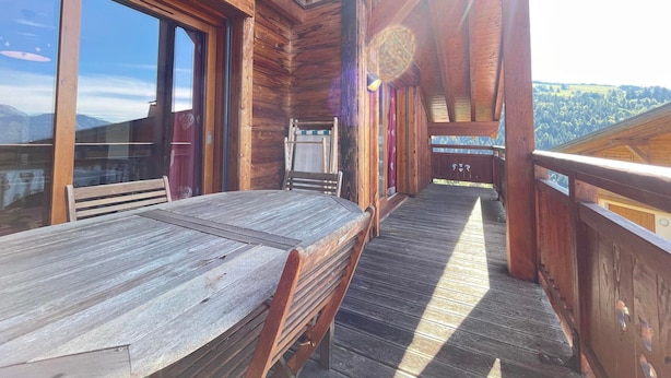 Una terraza de madera con una mesa y sillas, ventanas amplias y vistas al paisaje.