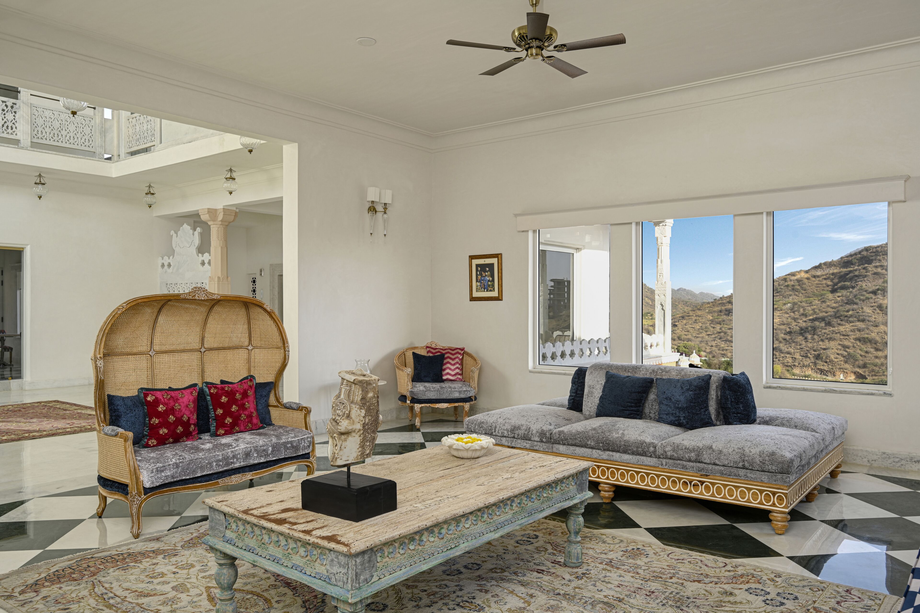 A spacious living room with a high ceiling, a large window offering a mountain view, a rattan sofa, a cushioned armchair, and a coffee table.