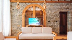 A stone-walled room with a wooden-framed TV stand, a sofa, and hanging light fixtures. - Kshira Oasis Qeparo - Boutique Beachfront Suites & Restaurant (Himare)