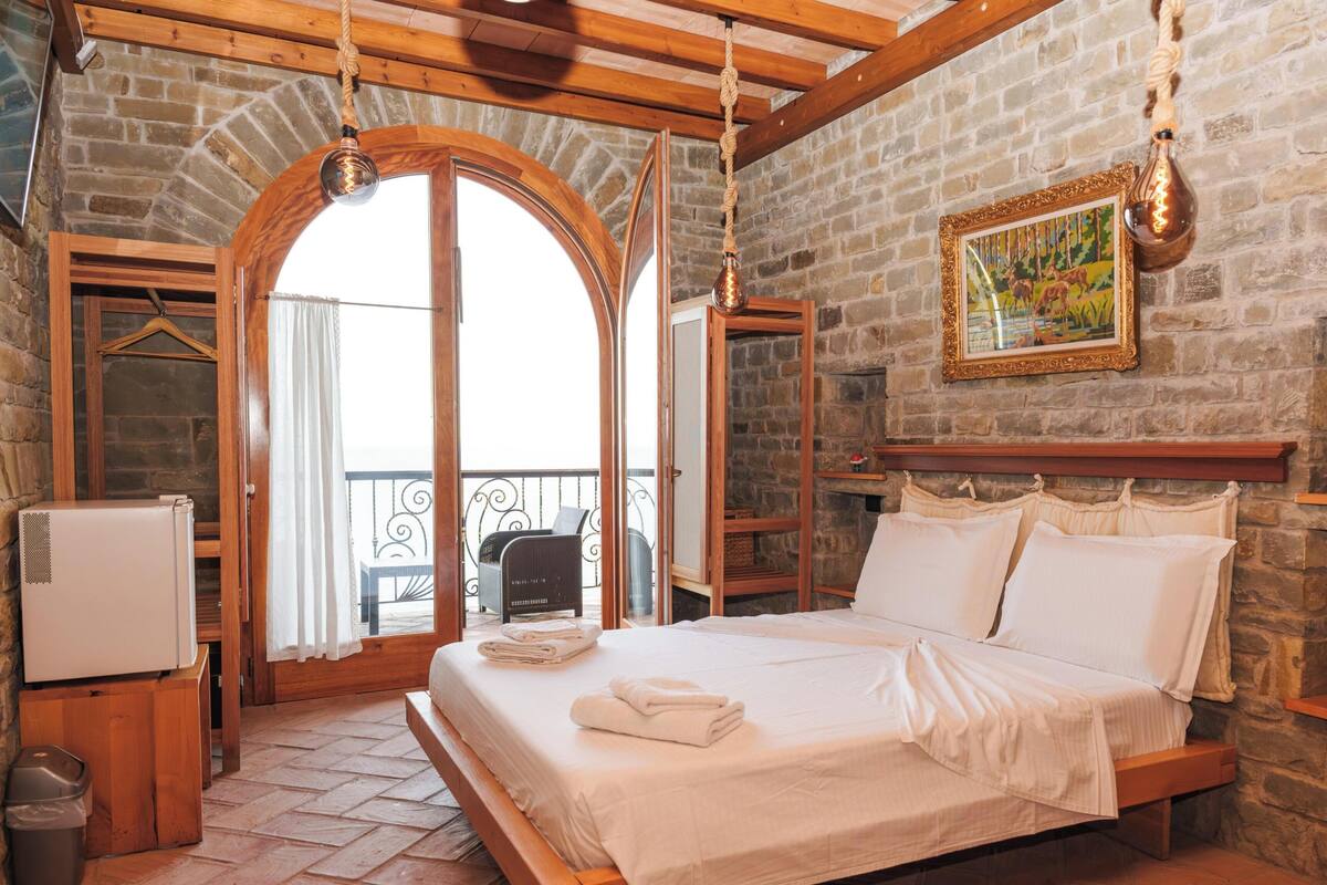 Una habitación con cama, ventana de madera, pared de piedra y una lámpara colgante.