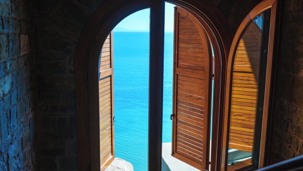 A view through an arched window with wooden shutters, overlooking a body of water. - Kshira Oasis Qeparo - Boutique Beachfront Suites & Restaurant (Himare)