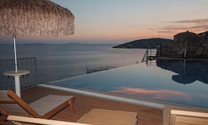Une piscine à débordement offrant une vue sur la mer, une terrasse en bois, des chaises longues et un parasol en chaume.