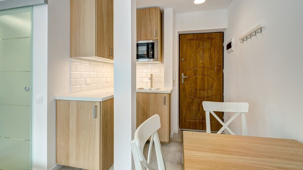 A compact kitchen with wooden cabinets, a microwave, and a white chair. - Mini Studio 2 by Grand Apartments (Sopot)