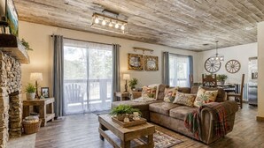 A rustic living room with a wooden ceiling, a stone wall, a sofa with decorative pillows, a wooden coffee table, and a dining area with a table and chairs. - Arcadian Pass, “Cozy Cabin Retreat in the Smokies – Comfort & Fun for All” (Sevierville)