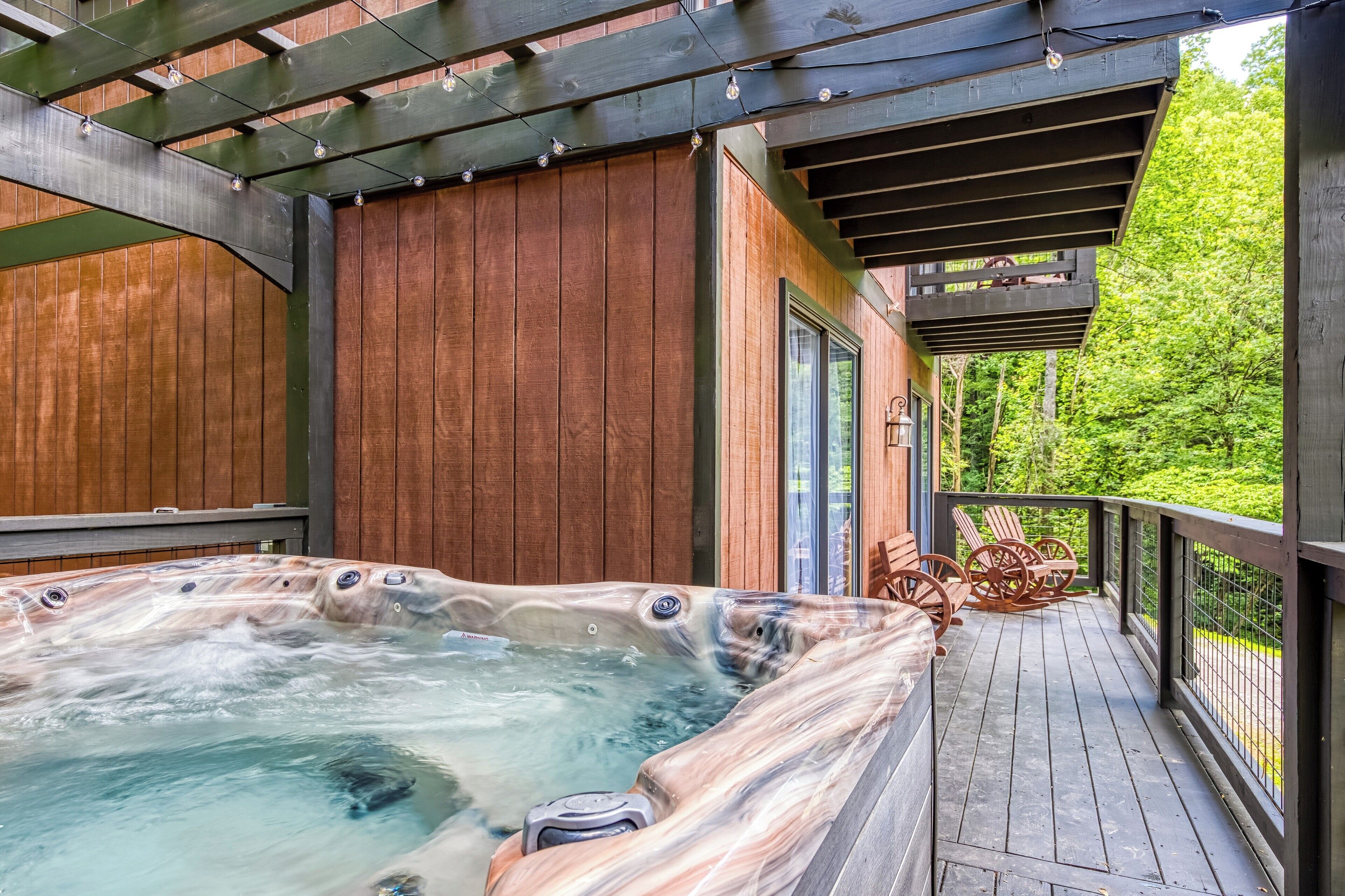 A hot tub on a wooden deck with a pergola overhead, surrounded by wooden walls and a view of greenery.