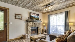 A cozy living room with a stone fireplace, wooden furniture, and a large window with curtains. - Arcadian Pass, “Cozy Cabin Retreat in the Smokies – Comfort & Fun for All” (Sevierville)