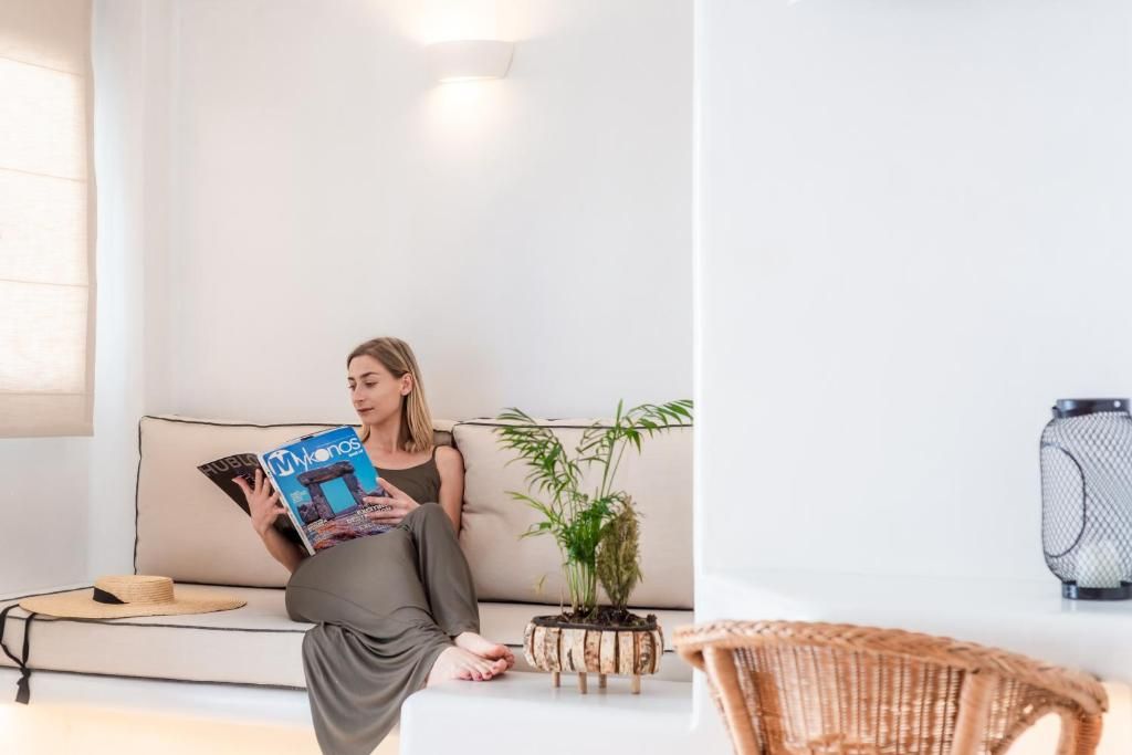 Una persona sentada en un sofá con una revista, una planta y una silla de mimbre en una habitación con pared blanca y una lámpara.