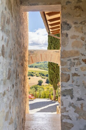 Un couloir en pierre mène à une arche en bois d’où l’on peut admirer un paysage verdoyant et une chaise rouge.