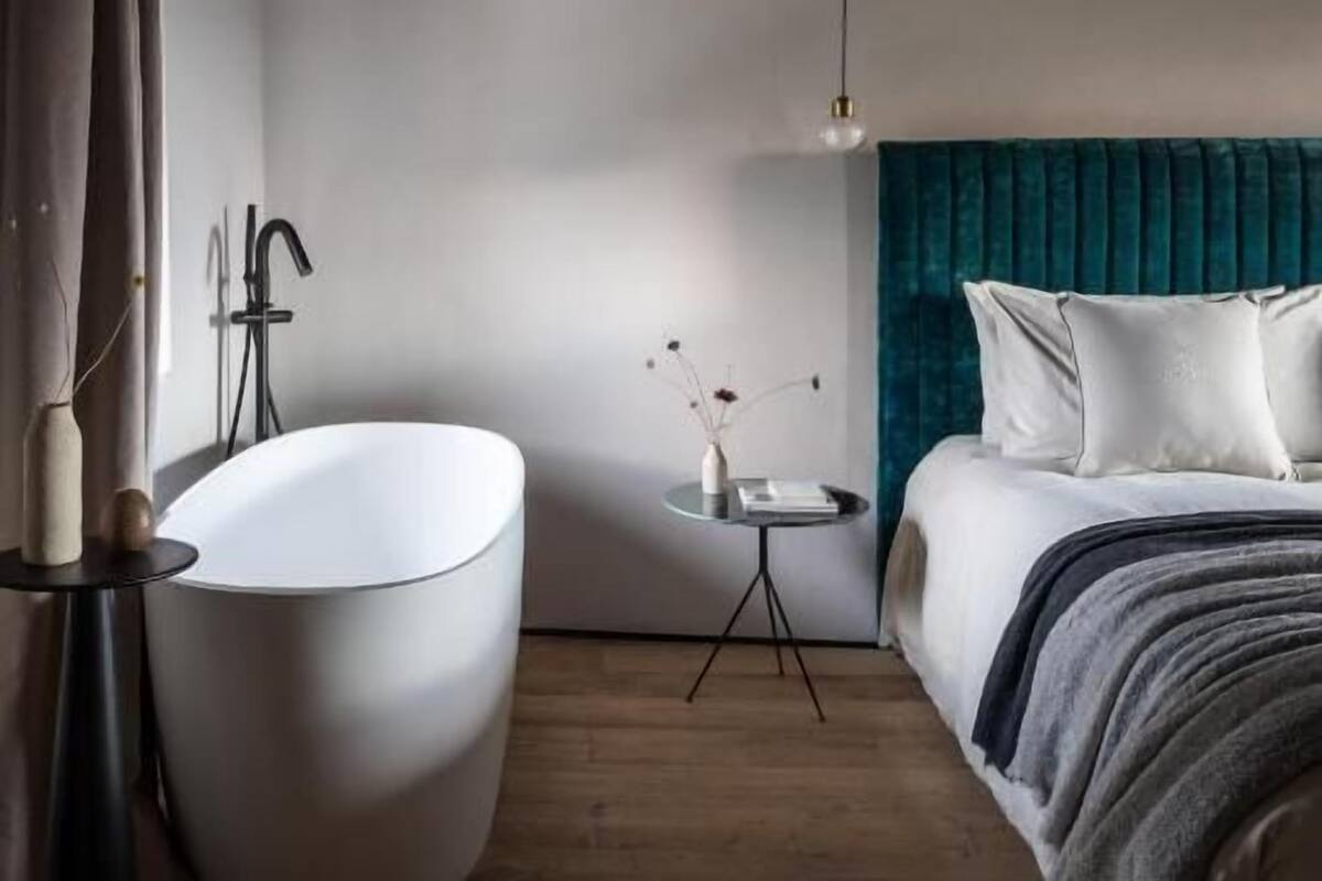 A modern bedroom with a large freestanding bathtub, a bed with teal bedding, and a small bedside table with a vase.