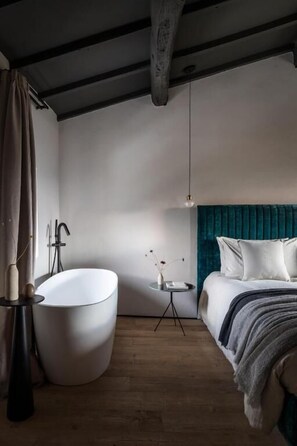 A modern bedroom with a large freestanding bathtub, a bed with teal bedding, and a small bedside table with a vase.