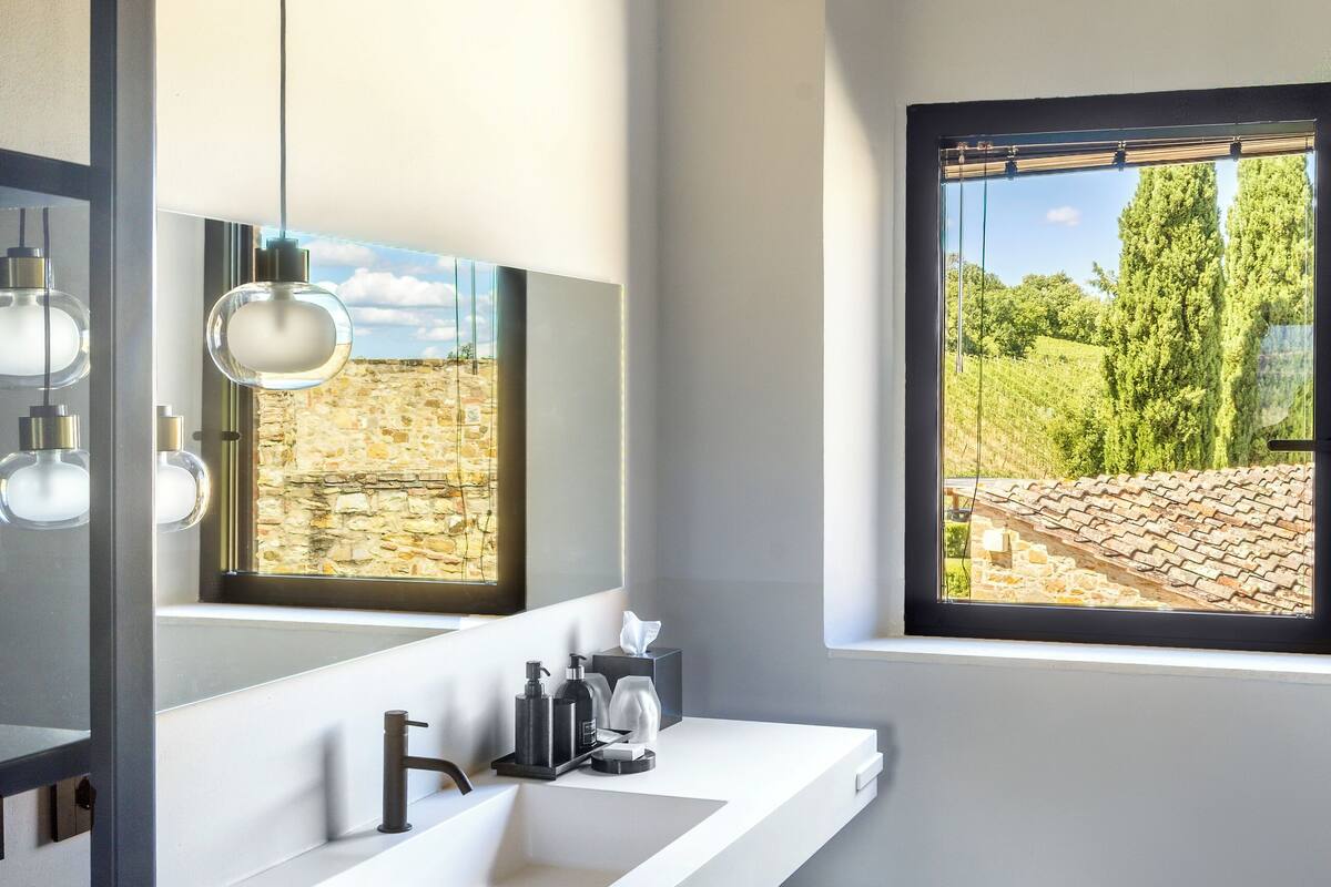 A modern bathroom with a white sink, blue cabinet, and a large mirror reflecting a stone wall and window.