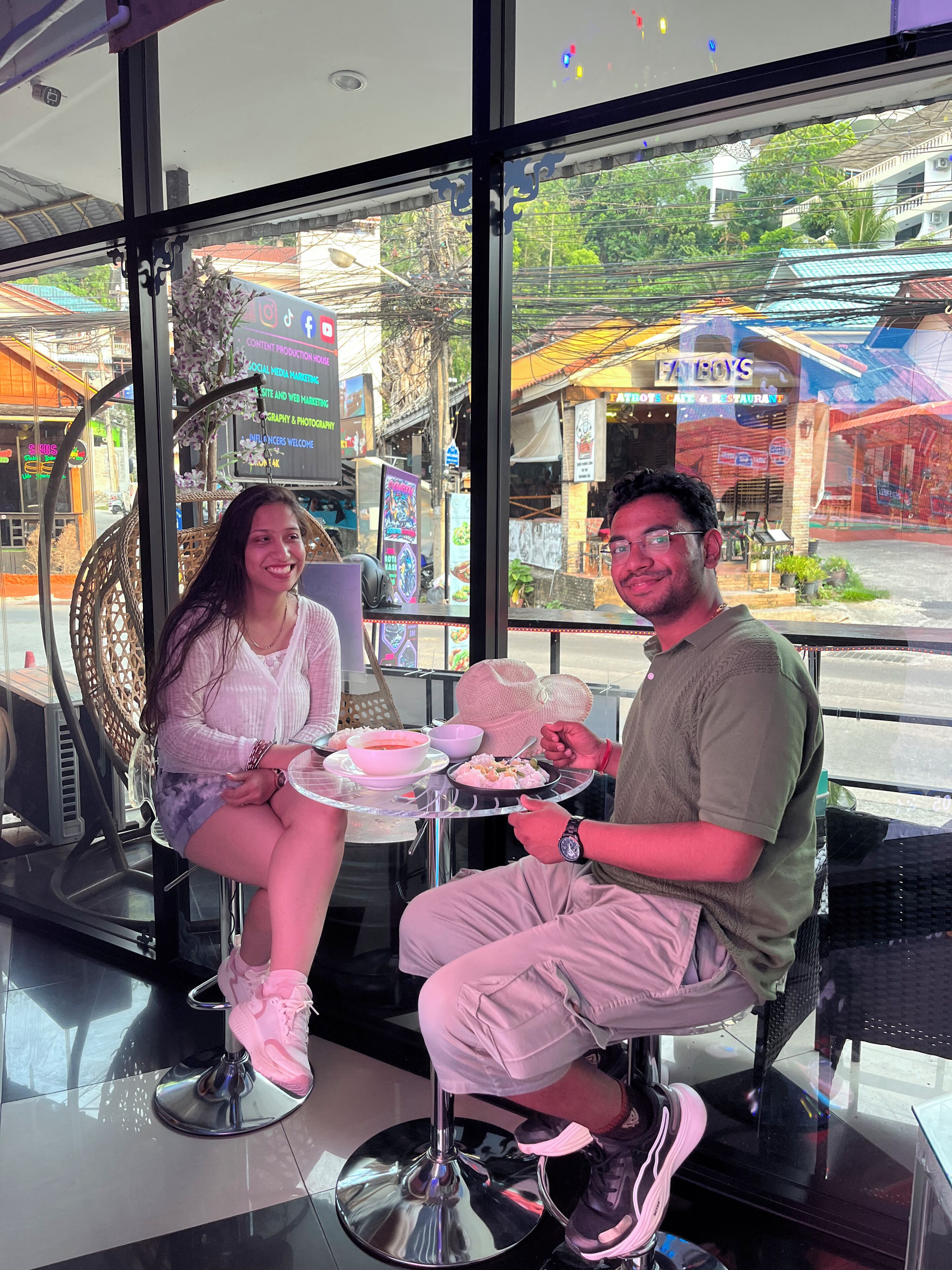 Two people seated at a table inside a building with large windows, a chair, and a small table with food.