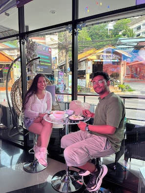 Two people seated at a table inside a building with large windows, a chair, and a small table with food.