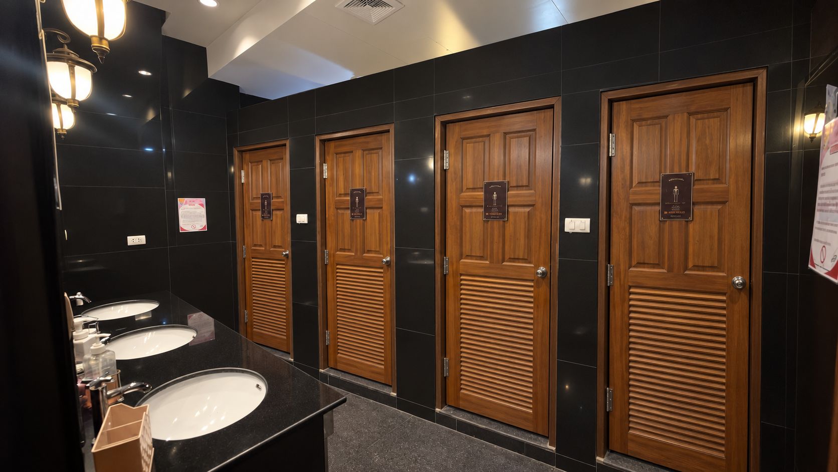 A public restroom with three sinks, wooden doors, and a black tiled wall.