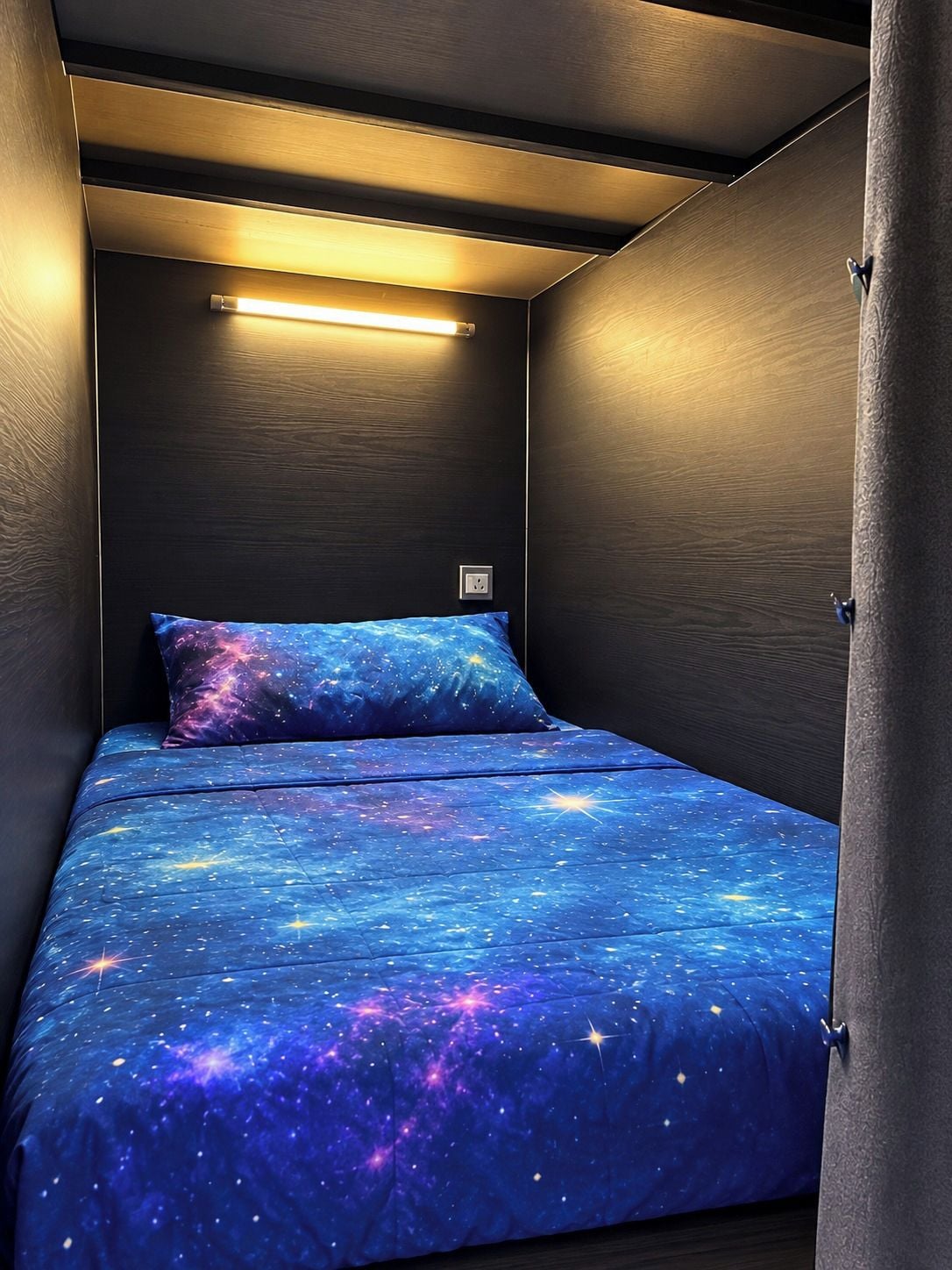 Ein kleines Zimmer mit einem Bett, das eine Bettdecke mit Galaxie-Motiv, einer Wandleuchte und einer Steckdose hat.