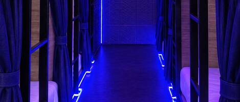 Un corridoio con letti a castello, ognuno dotato di tenda, e una striscia luminosa al neon blu sul pavimento.